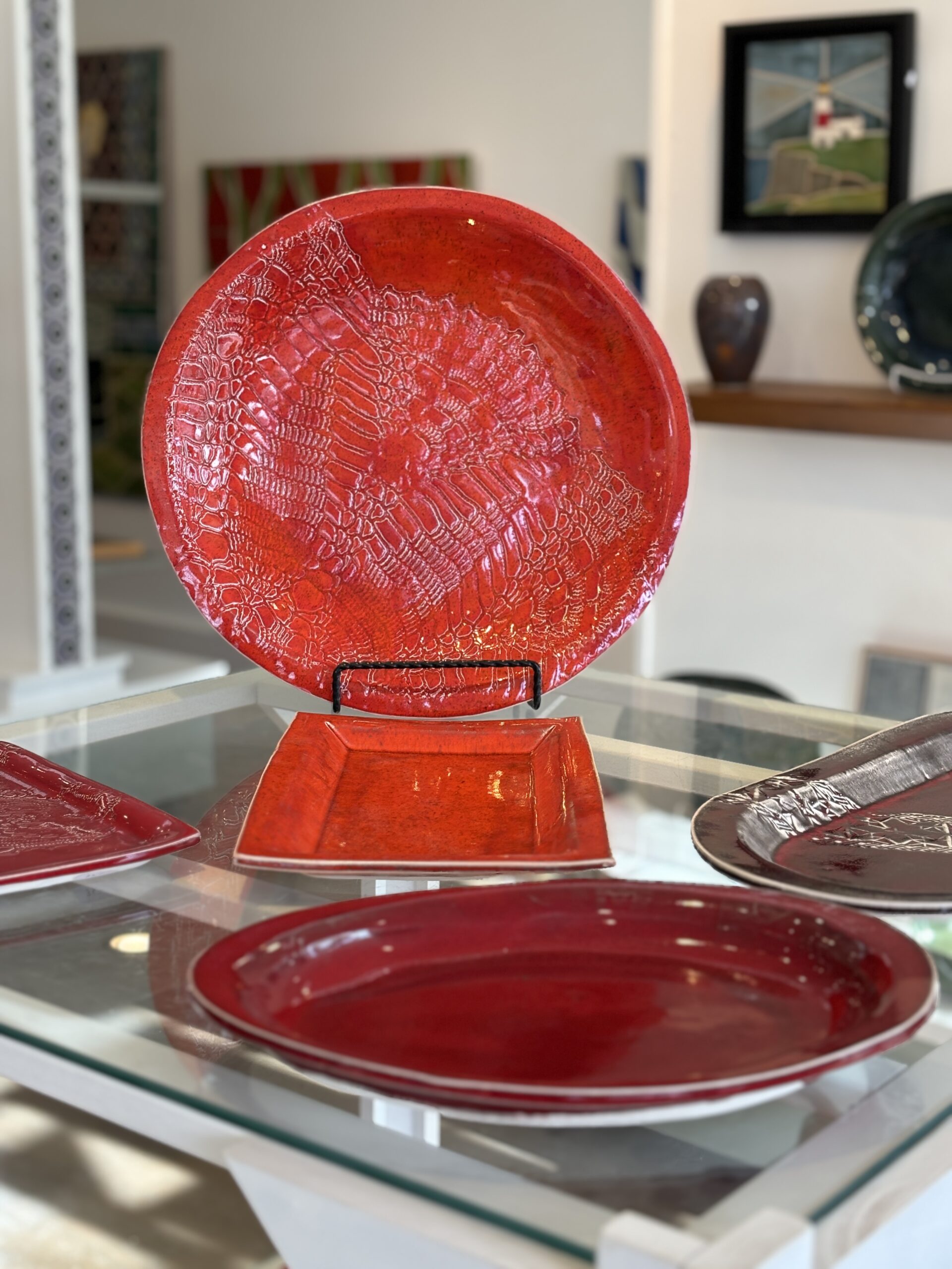 A red dish with a fossil like tecture displayed on a glass table top in front of other red dishes.