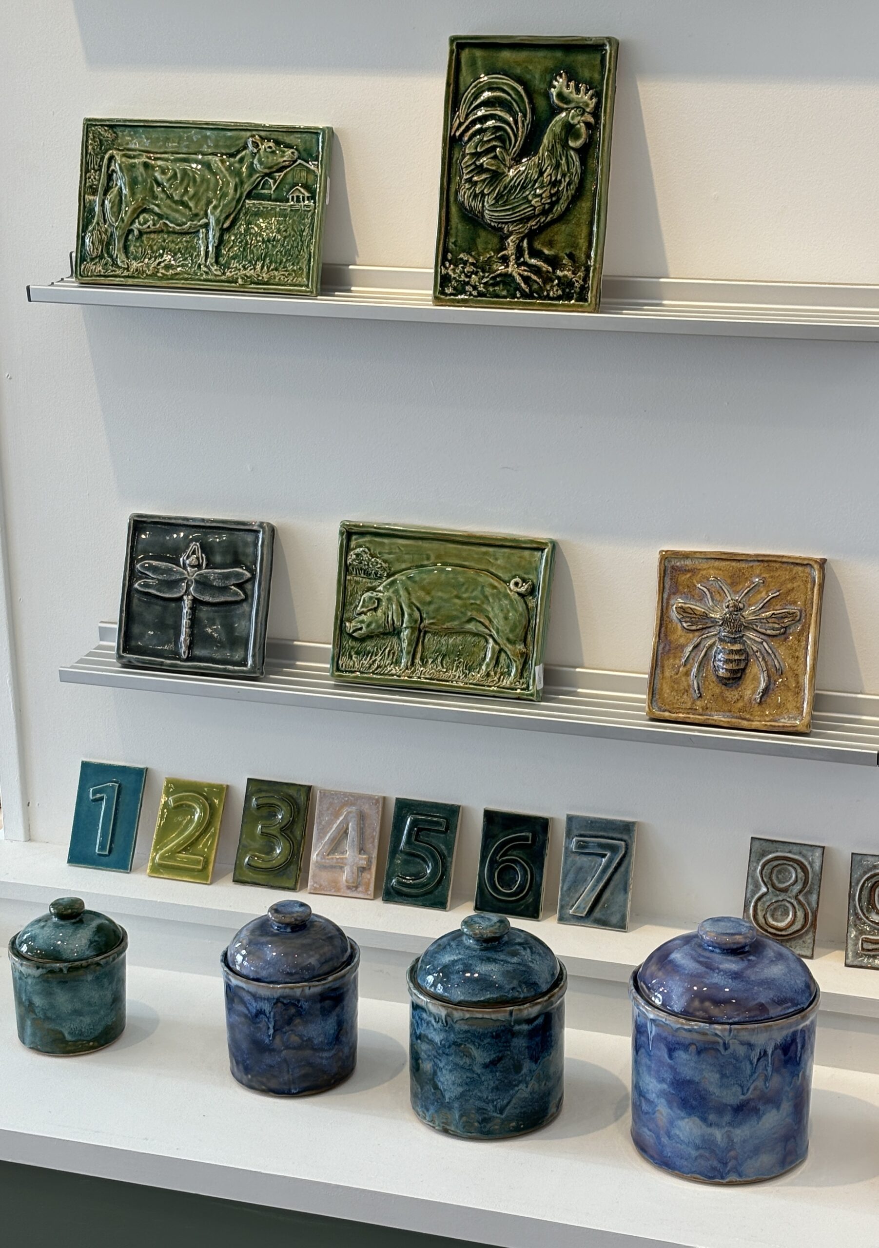 A wall of various colored ceramic tiles imprinted with animals and bugs. Below are various colored tiles imprinted 1 through 9. In front of them are four blue colored ceramic pots with tops.