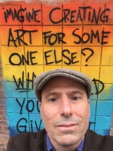 A person in a hat in front of a painted wall that says Imagine Creating Art For Someone Else?