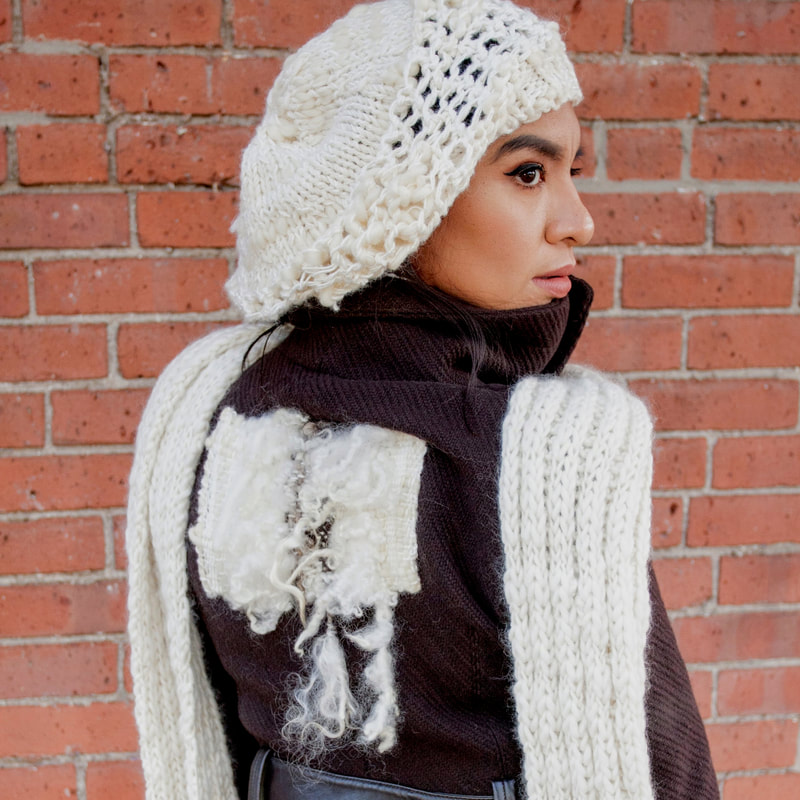 A woman looking over her shoulder with a hand knit white hat, scarf, and wool design on the back of her shirt.